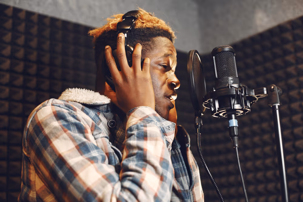 Young man wearing headphones and a plaid jacket singing into a microphone in a soundproof recording studio.