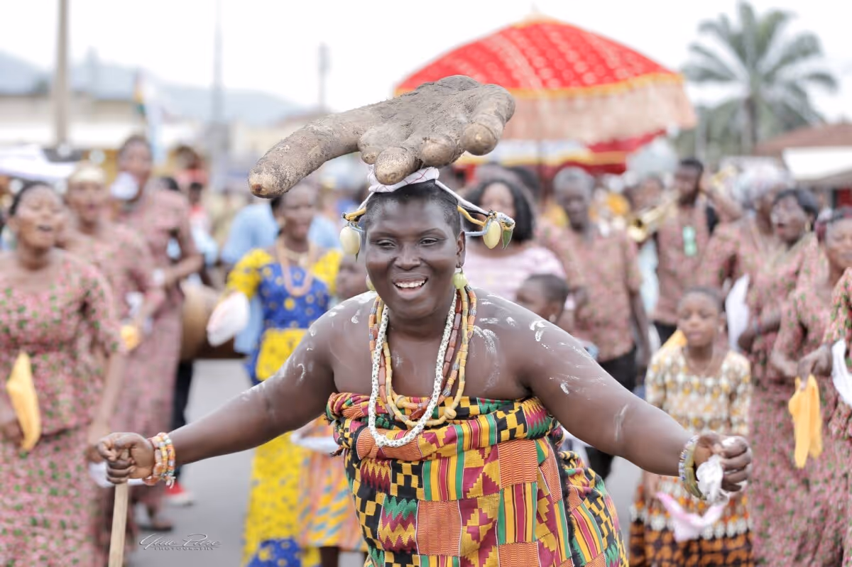 Smiling woman in colorful traditional clothing and beads dancing outdoors with a yam tied on her head, surrounded by people in festive attire.