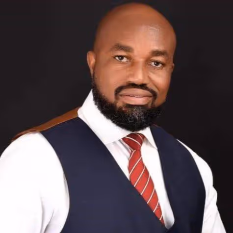 Portrait of a bald man with a beard wearing a white shirt, navy blue vest, and red striped tie against a black background.