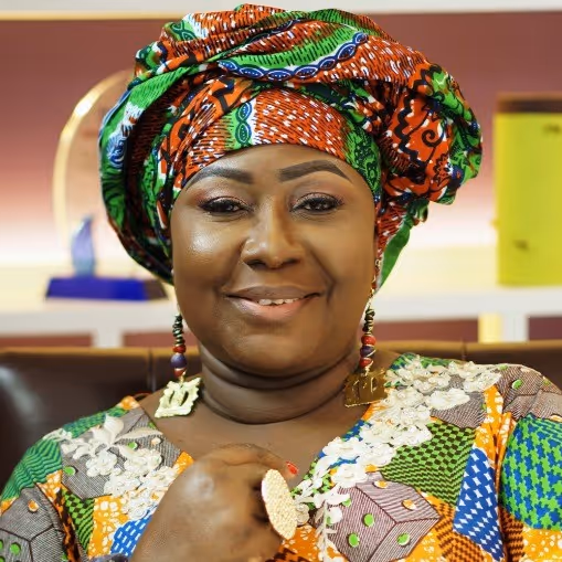 Smiling woman wearing colorful patterned traditional African attire and matching headwrap, seated indoors.