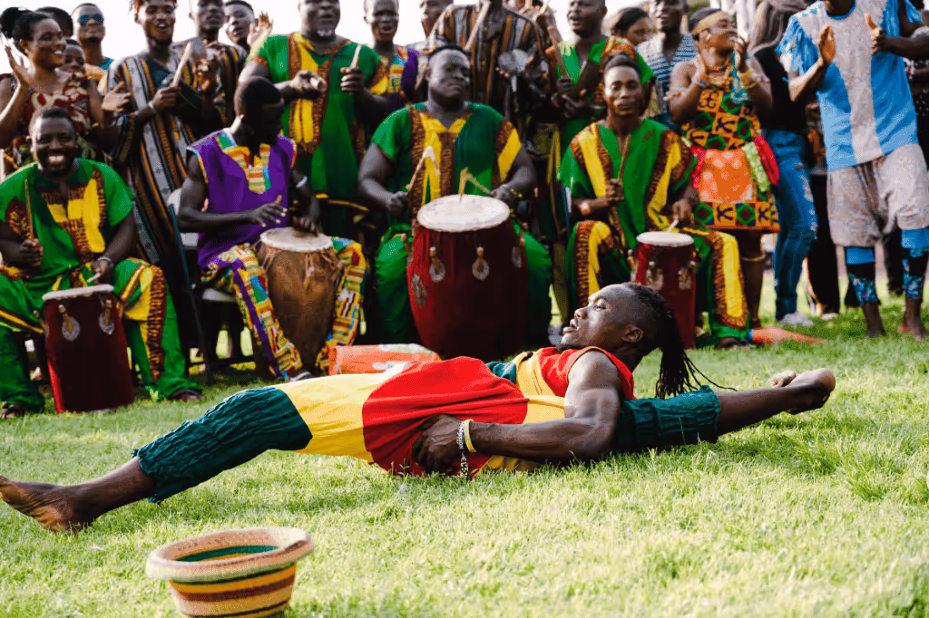 A male dancer in colorful traditional attire performing a ground-level dance move while musicians in vibrant clothing play drums behind him outdoors.