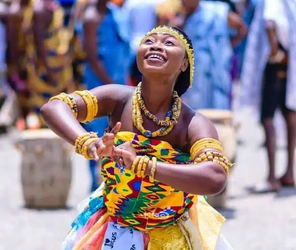 Smiling woman in colorful traditional Ghanaian attire and beads performing a cultural dance outdoors.