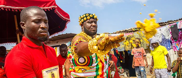 Man dressed in colorful traditional attire throws maize during a cultural festival, surrounded by onlookers.