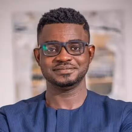 Portrait of a smiling man with short curly hair, glasses, and a blue shirt, standing indoors.