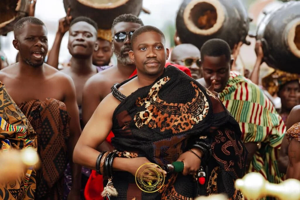 A young man dressed in traditional African attire stands confidently among a group of similarly dressed men during a cultural ceremony.