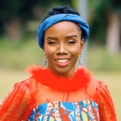 Smiling woman wearing a blue headband, red feathered collar, and patterned clothing outdoors.