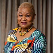 Portrait of an elderly woman with short blonde hair wearing a colorful patterned dress and beaded necklace, smiling at the camera.