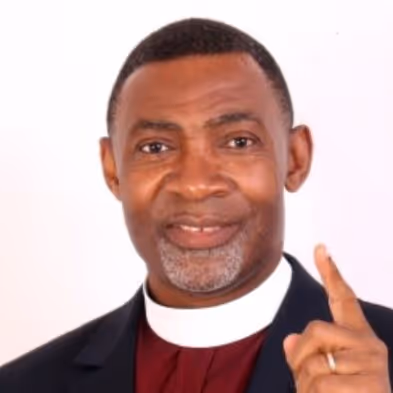 Smiling man wearing clerical collar and dark jacket, raising his right index finger.