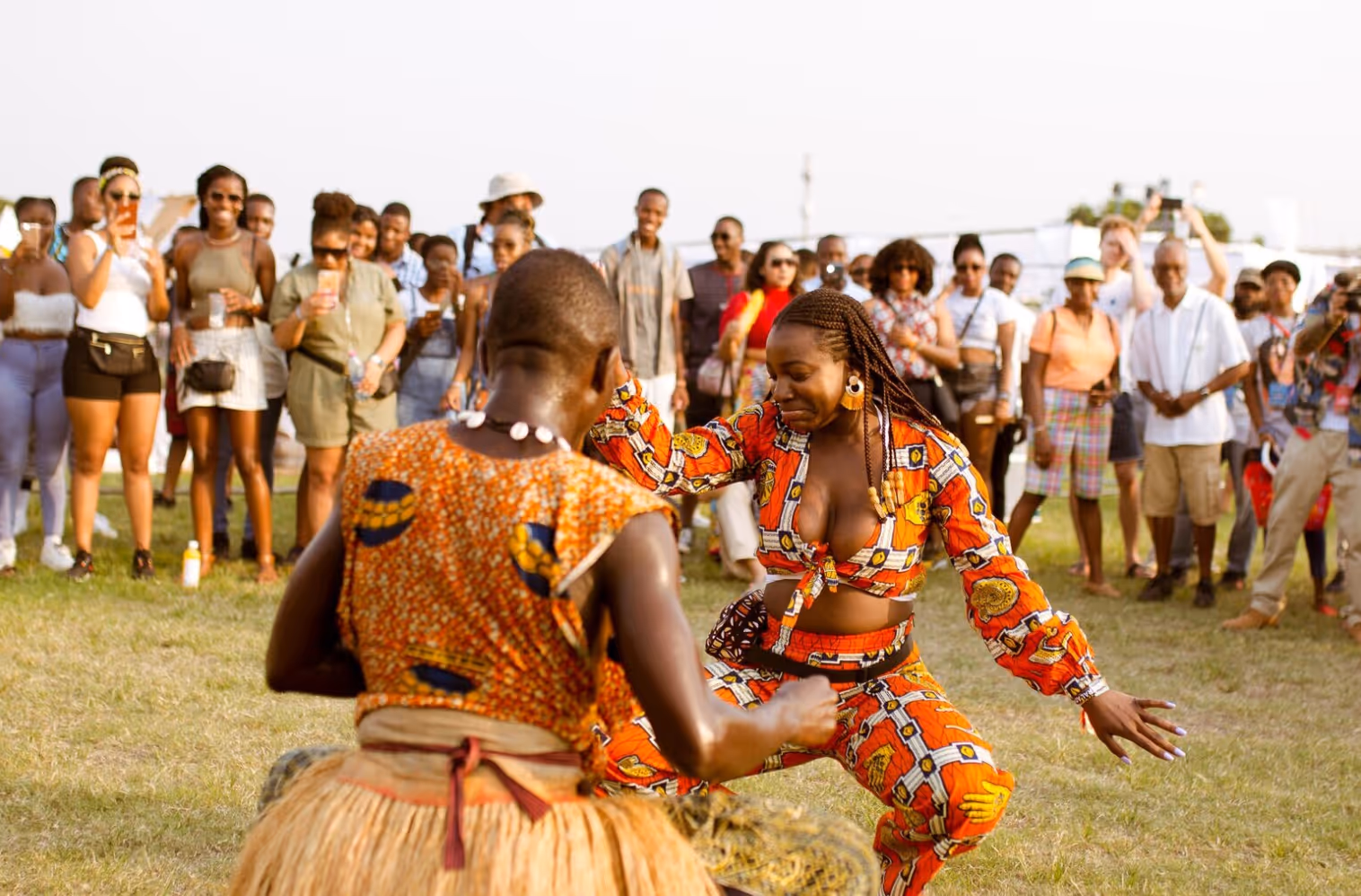 Two people dressed in vibrant traditional African attire dancing outdoors with a crowd watching in the background.
