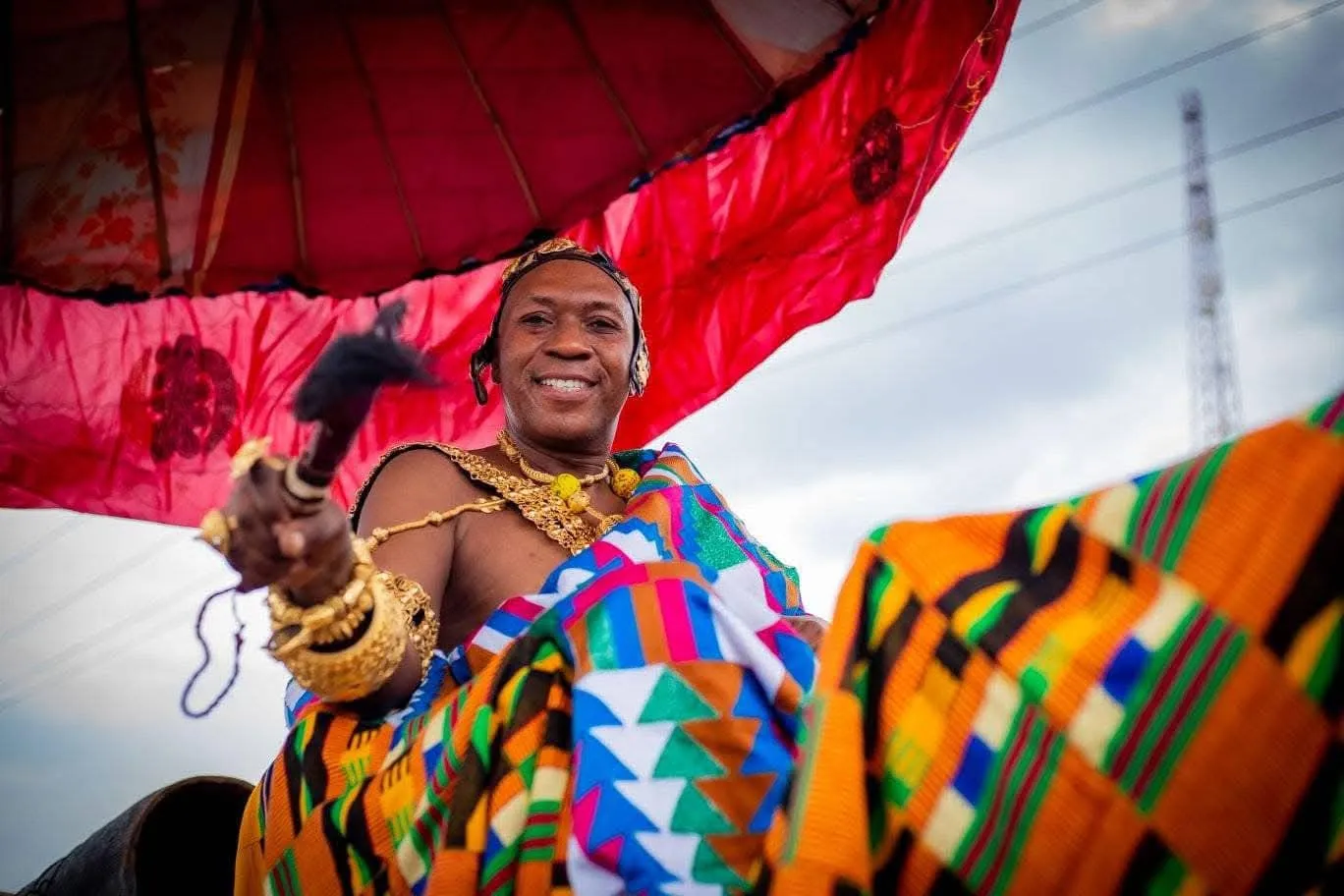 Smiling man dressed in colorful traditional African attire and gold jewelry holding a staff under a large red umbrella.