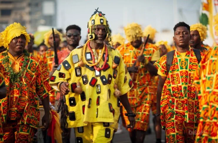 Group of men wearing colorful traditional African attire with one man in the center wearing a yellow outfit decorated with black and red patterns and beads.