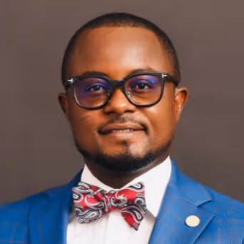 Man wearing a blue suit, white shirt, patterned red and white bow tie, and dark-rimmed glasses against a plain brown background.