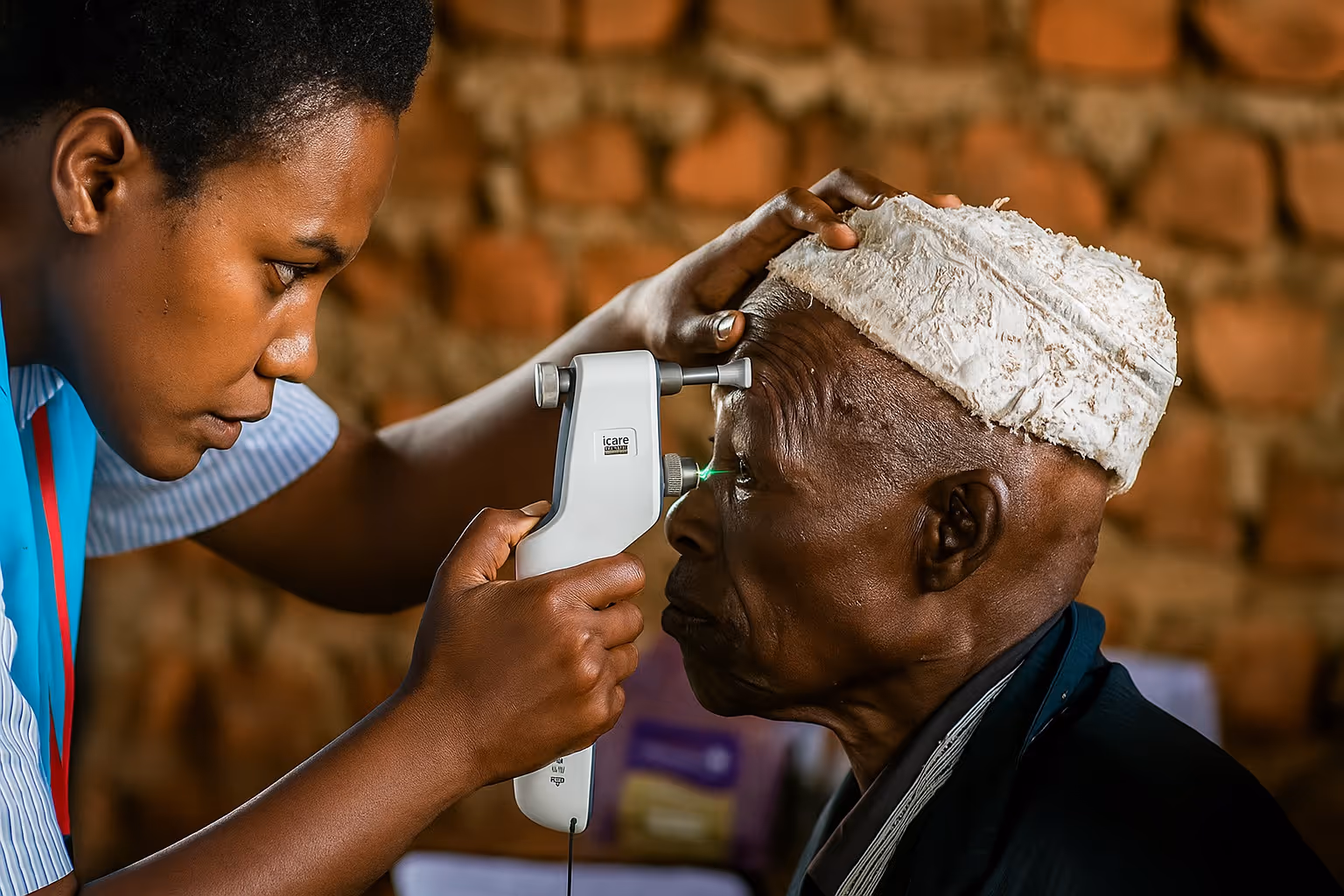 Healthcare worker using an ophthalmic device to examine an elderly man's eye in a brick-walled setting.