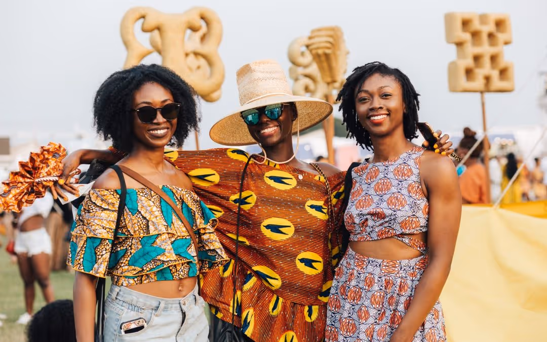 Three smiling women wearing colorful African print clothing at an outdoor event with artistic sculptures in the background.