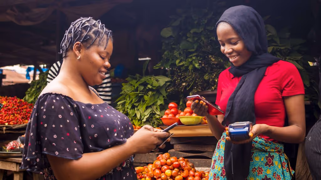 Two women smiling and using mobile phones for a payment transaction at an outdoor market with tomatoes and greens in the background.