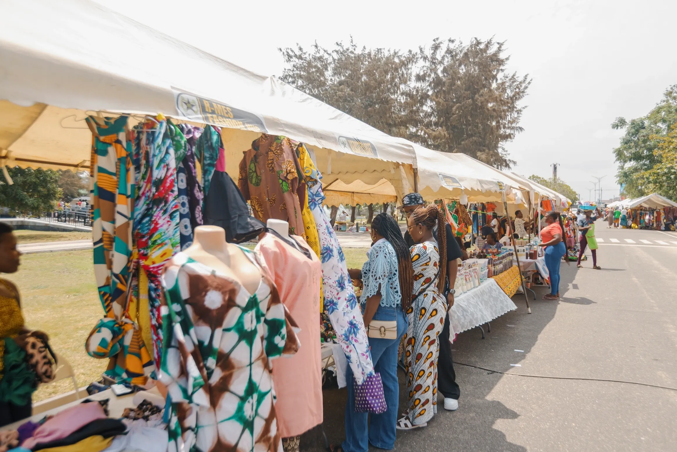 Outdoor market stalls with colorful fabrics, clothes, and handmade goods displayed, and people browsing under white tents on a sunny day.